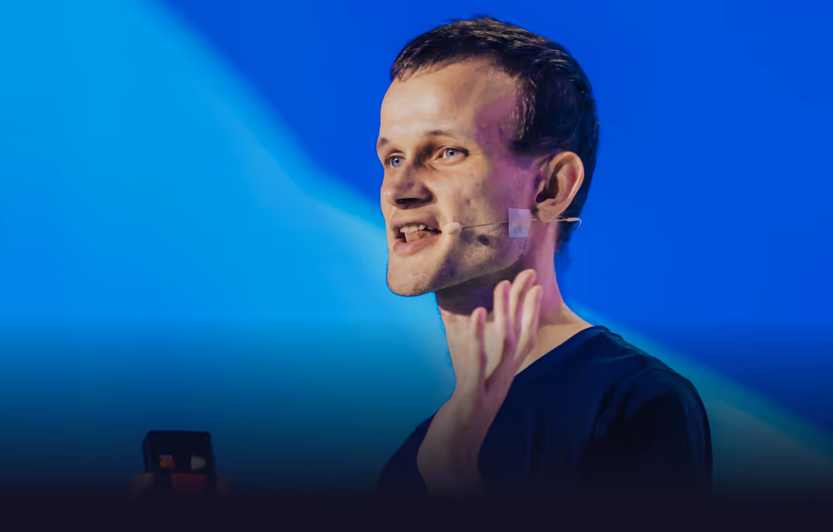Man speaking on stage with a headset microphone against a blue background, holding a smartphone in one hand and gesturing with the other.