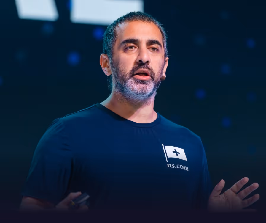 Man with short dark hair and beard speaking on stage wearing a black t-shirt with a small flag and 'ns.com' logo.